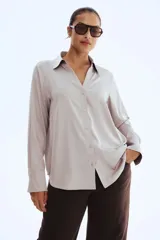 Blusa de manga larga color gris claro o beige pálido, con acabado satinado y cuello clásico que forma un escote en V profundo con cierre de botones frontales.