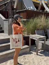 Tote bag de tela color crudo con asas blancas y estampado de un bikini secándose en una cuerda.