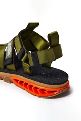 Sandalias Kenner Rakka Siren de tiras de tela verde militar, con ajuste de velcro en el tobillo y suela de goma marrón con detalles en naranja.
