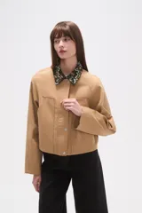 Campera corta color beige, con cuello camisero estampado floral en tonos oscuros y cierre frontal de botones a presión. Presenta bolsillos superiores con solapa y pespuntes verticales en el frente.