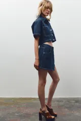 Falda pantalón corta de tiro alto confeccionada en denim azul oscuro. Presenta bolsillos delanteros y traseros de parche. El cierre es cruzado con cremallera oculta y botón metálico.
