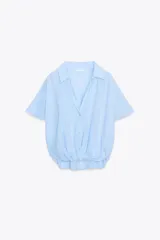 Camisa color gris claro con cuello solapa y escote pico, de manga corta. Presenta un nudo en la parte delantera y cierre frontal con botones.