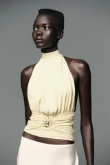 Top color crema con cuello halter, espalda descubierta, detalle de frunces y pieza metálica en forma de sol en el delantero.
