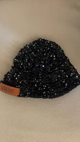 Casquete tejido a mano color negro, con un patrón abierto y adornado con lentejuelas brillantes. Posee una etiqueta de cuero marrón con el nombre "Soriame" grabado.