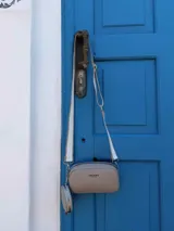 Bandolera pequeña color negro, de material símil cuero, con forma rectangular y cierre superior. Incluye un monedero redondo desmontable con cierre, ambos con logo grabado en blanco. La correa es ancha y de tela con diseño a rayas en blanco y negro.