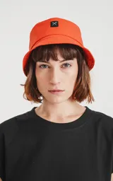 Gorro tipo bucket de tela color naranja vibrante, con un pequeño parche negro en el frente que tiene un logo bordado en forma de tijeras cruzadas.