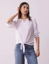Blusa blanca de tejido texturado con cuello redondo, escote en V con aplique metálico y mangas 3/4 con puños elásticos. El ruedo se ata con un nudo.