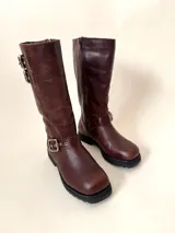 Bota de caña media de cuero vacuno color marrón oscuro, con punta cuadrada, cierre lateral y suela track negra. Presenta detalles de hebillas metálicas plateadas en la caña y en la parte inferior del empeine.