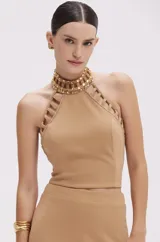 Top cropped color beige confeccionado en punto crepé, con cuello halter adornado con apliques dorados y aberturas caladas en los hombros.