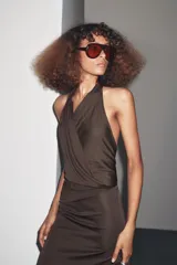 Vestido largo color marrón oscuro, de cuello halter y escote fluido. Tiene hombros descubiertos, detalle de pliegues laterales, forro interior a tono y cierre lateral con cremallera oculta y en espalda con botones.