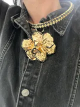 Collar con cadena gruesa tejida en cuero negro y dorado, con dije grande dorado en forma de flor con borde negro.