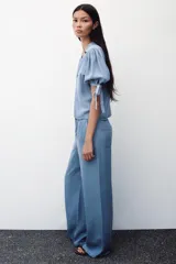 Camisa celeste de denim con cuello solapa, escote en pico, mangas abullonadas con lazos y cierre frontal con botones.