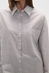 Camisa de algodón blanca de corte holgado, con cuello camisa y manga larga. Presenta un estampado de finas rayas verticales negras y un bolsillo frontal con rayas horizontales.