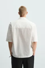 Camisa de corte relajado en color blanco, confeccionada en tejido de algodón con textura de aplicaciones. Posee cuello bowling, manga corta y cierre frontal con botones.