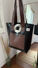 Bolso tote de ecocuero marrón oscuro con textura cocodrilo y un parche frontal de cuero liso marrón claro. Posee asas de hombro de cuero marrón y un broche decorativo blanco en el frente.