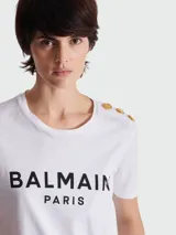Remera blanca de algodón con cuello redondo y mangas cortas. Estampado frontal con el logo 'BALMAIN PARIS' en color negro. Detalle de botones dorados en el hombro izquierdo.