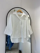 Camisa blanca de manga larga con cuello camisero y cierre frontal con botones. Presenta bordados perforados combinados a tono. El bajo tiene aberturas laterales y la parte trasera es más larga que la delantera.