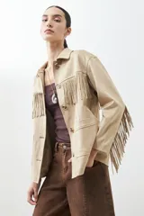 Chaqueta color beige de simil gamuza con flecos en mangas y ruedo, con cuello camisero y cierre frontal con botones.