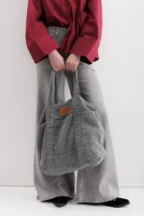 Bolso tote grande de jean gris con asas de mano y asa larga para colgar. Se cierra con un botón de metal a presión y contiene tres bolsillos exteriores.
