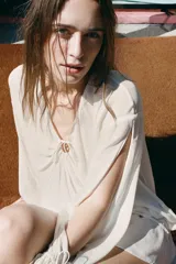 Top semitransparente color crema, de escote pico ajustable con lazadas, manga larga con aberturas y puño ajustable con cordones.