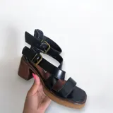 Sandalias de cuero negro con tiras anchas, hebillas doradas y tacón de madera.