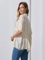 Blusa color crema de crepe con cuello a la base con solapa, escote en V con abertura central y botones. Delantera con detalles plisados y apliques en crochet. Manga corta con terminación fruncida y ruedo en broderie.
