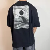 Remera negra de corte oversize con estampa rectangular en la espalda que representa una mano alcanzando un círculo sobre un fondo de líneas horizontales.