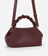 Cartera pequeña color borgoña, con asa corta trenzada y correa larga ajustable y desmontable. Cierre superior con broche imantado. Interior forrado con bolsillo.