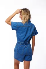 Mono corto de jean azul con cuello camisero, cierre frontal con botones, mangas cortas con dobladillo y bolsillos delanteros y traseros.