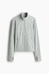 Campera deportiva color gris claro, de corte ajustado, con cuello elevado, cierre frontal, bolsillos laterales con cierre y mangas largas con abertura para el pulgar.