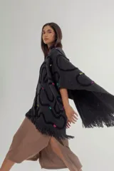 Ruana de tejido de punto grueso color gris oscuro con flecos en el borde inferior. Presenta apliques de lana en forma de líneas onduladas en color negro, fucsia, amarillo y celeste.