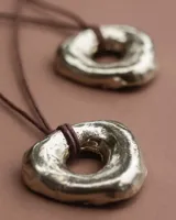 Collar con dije dorado irregular con agujero central, colgando de cordón negro ajustable.