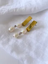 Aros colgantes con base rectangular dorada y dos perlas blancas unidas por aros de metal.