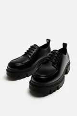 Zapato de vestir tipo blucher color negro, con detalle de bordón, tirador trasero y horma redonda. Suela con volumen y diseño irregular.