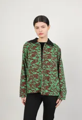 Campera de jean con cuello clásico, cierre frontal con botones metálicos, dos bolsillos de parche en el pecho y estampado floral en tonos verdes y marrones.