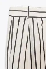 Pantalón de tiro medio con estampado de rayas verticales negras sobre fondo blanco. Tiene trabillas en la cintura, bolsillos laterales, pinzas delanteras y cierre frontal con cremallera, botón y ganchos metálicos.