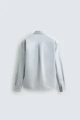 Camisa celeste a rayas verticales blancas, de corte relaxed fit, cuello solapa, manga larga con puños abotonados, bolsillo de parche en el pecho y cierre frontal con botones.