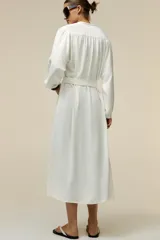 Vestido midi blanco de crepe con cuello redondo y escote pico, manga larga con puños y cinturón de la misma tela con hebilla metálica. Cierre frontal con botones metálicos.