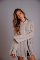 Camisa color beige claro, de corte holgado, con cuello clásico, cierre frontal con botones, mangas largas con puños abotonados y detalles de broderie en la parte delantera.