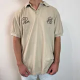 Chomba color beige con cuello clásico, corte oversize, mangas cortas y bordados marrones con la inscripción "Golf Club" y otros motivos. 