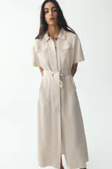 Vestido camisero largo color beige, de manga corta y cuello solapa. Tiene bolsillos de parche en el pecho, bolsillos laterales ocultos y cinturón de la misma tela.