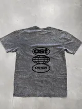 Remera gris con efecto acid wash, corte oversize y estampado trasero en negro con el texto "ost" en un óvalo, un diseño de globo terráqueo con líneas de latitud y longitud, y el texto "osten" en otro óvalo.