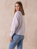 Blusa de algodón a rayas verticales finas en tonos lila y blanco, marca Dash. Escote en V y abertura frontal con botones. Mangas largas regulables con presilla y botón. Terminación de ruedo anudado.