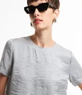 Blusa plateada de mangas cortas con cuello redondo pespuntado, botón en la espalda y superficie metalizada.