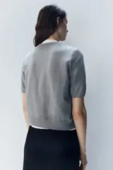 Cárdigan gris de punto fino con cuello redondo, manga corta y cierre frontal con botones. Tiene terminaciones acanaladas en cuello, puños y ruedo.