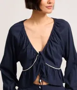 Blusa de mangas largas color azul marino, confeccionada en viscolino con 30% de lino, con escote V, lazo en el cuello con punta y bieses contrastantes luego abajo del escote, además del bajo de volado.