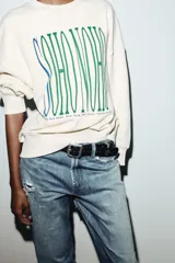 Sudadera de tejido mezcla de algodón color crema, con cuello redondo y manga larga. Presenta un estampado central con el texto "SOHO NOHO" en tipografía alargada, con las letras alternando entre azul y verde.