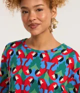 Remera de manga corta con cuello redondo y estampado repetitivo de loros rojos y hojas verdes sobre fondo azul y rojo. El borde de las mangas y el cuello es de punto acanalado verde.