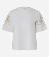 Remera blanca de algodón con corte slim, cuello redondo y mangas cortas. Presenta un bordado lineal con motivo floral en tono dorado/marrón claro sobre los hombros y mangas.