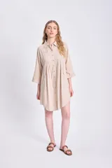 Vestido camisero corto color beige, con cuello y botones frontales. Presenta pliegues verticales (tablas) en el área del pecho y un corte holgado con ruedo asimétrico.